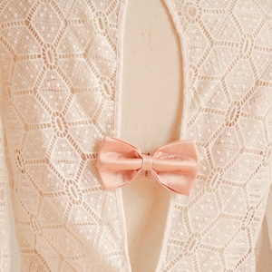 以下が含まれることがあります： 背中が大きく開いた白いレースのドレスに、ピンクのリボンが飾られています。ドレスは幾何学模様で、長袖です。