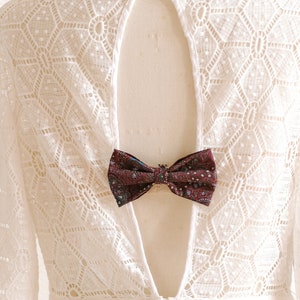 May include: White lace dress with a low back and a burgundy bow tie accent. The dress has a geometric pattern and is on a white mannequin.