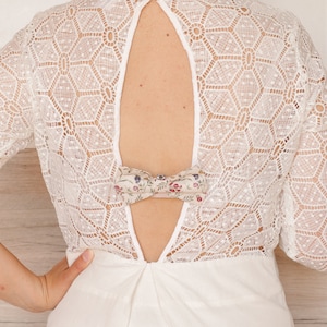 May include: White lace dress with a low back and a floral bow detail. The dress has a geometric pattern and a fitted silhouette.