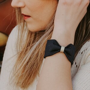 Könnte beinhalten: Eine Frau mit langen braunen Haaren trägt einen weißen Pullover und ein schwarzes Schleifenarmband mit goldener Verzierung.