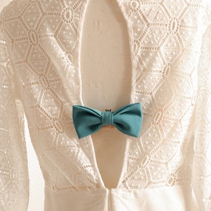 May include: White lace dress with a low back and a teal bow tie accent. The dress has a geometric pattern and is on a white mannequin.
