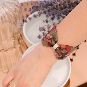 May include: A green, red and brown patterned fabric bracelet with a gold metal accent. The bracelet is tied in a bow on a person's wrist.