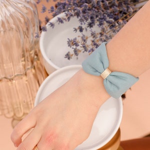 Puede incluir: Una pulsera de tela azul claro con un lazo dorado. La pulsera está atada a la muñeca de una persona.