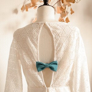 May include: White lace dress with a low back and a teal bow tie accent. The dress has a geometric pattern and is on a white mannequin.