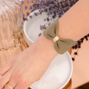 May include: A green fabric wrist cuff with a gold bow. The cuff is tied in a bow and is worn on a person's wrist.