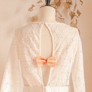 May include: A white lace dress with a low back and a peach-colored bow tie. The dress has a geometric pattern and long sleeves.