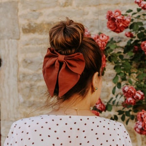 Peut inclure: Un grand nœud pour cheveux en tissu de couleur rouille unie fixé sur un élastique, porté dans un chignon haut. L'accessoire est associé à un vêtement de couleur claire orné de petits pois marron foncé et à un fin collier doré.
