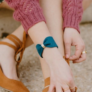 May include: A woman wearing a pink sweater and a floral skirt is tying the laces of a brown suede heeled shoe. She is wearing a blue ribbon bracelet on her wrist.