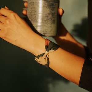 May include: A fabric bracelet with a bow design. The bracelet has a mix of black, tan, and red colours, and is secured with a gold-coloured band. A grey mug is held in the background.
