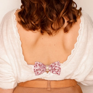 Puede incluir: Un suéter de punto blanco con un lazo de estampado floral atado en la espalda. El suéter tiene la espalda baja y un cinturón marrón.