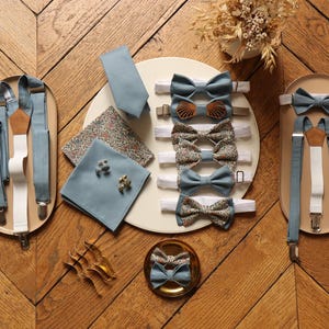 May include: A set of blue bow ties and suspenders with a floral patterned pocket square and cufflinks. The bow ties are arranged in a circle on a white plate, and the suspenders are on a wooden surface.