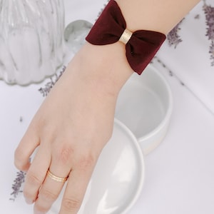 May include: A burgundy bow cuff bracelet with a gold centre detail. The bow is made of a soft fabric and is worn on the wrist. The bracelet is a fashion accessory and is shown on a person's arm.
