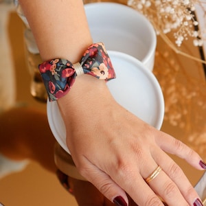 May include: A floral patterned fabric bow tied around a wrist. The bow is dark blue with red, pink, and white flowers. The bow is tied with a gold ribbon.