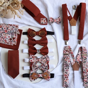 Op de afbeelding: Een set strikjes en bretels in een bordeauxrood en bloemenpatroon. De strikjes zijn gemaakt van verschillende stoffen, waaronder een strikje met een houtlook. De bretels zijn verstelbaar en hebben een leren band.