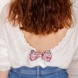 May include: A white knitted sweater with a floral bow on the back. The sweater has a low back and a lace trim. The bow is pink and white with a floral pattern.