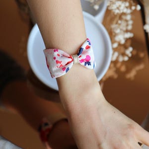May include: A floral bow bracelet with a white background and red, pink, and blue accents. The bow is tied in the center with a gold band. The bracelet is worn on a wrist.