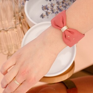 May include: A pink fabric wristband with a gold bow. The wristband is tied in a bow and has a gold accent.
