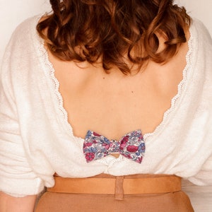 Puede incluir: Un suéter blanco con un lazo de estampado floral en la espalda. El suéter tiene la espalda baja y un cinturón marrón.