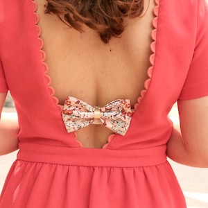 May include: Coral-colored dress with a bow detail. The dress features a low back with a scalloped edge and a floral bow. The short-sleeved dress has a fitted waist and a flowing skirt, suitable for special occasions.
