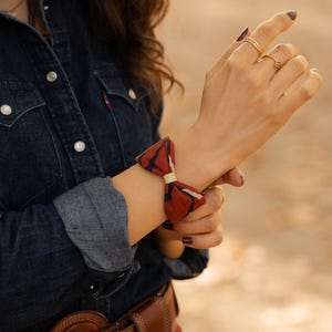 May include: A red, white, and navy blue patterned bow-tie bracelet. The bracelet is tied around a wrist, and the person is wearing a denim shirt and gold rings. A brown leather belt with a large buckle is also visible.