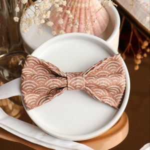 May include: A brown bow tie with a white wave pattern on a white plate. The bow tie is made of fabric and has a satin finish.