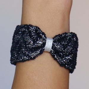 May include: A black bow bracelet with a silver band. The bow is made of a textured fabric with iridescent threads. The bracelet is worn on a wrist.