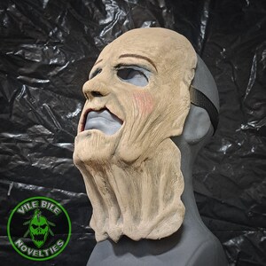 May include: A detailed, realistic, beige-colored, latex horror mask with a wrinkled, aged appearance. The mask features a wide, open mouth with a gray painted tongue and a furrowed brow. The mask is designed to be worn over the head and has a black elastic strap.