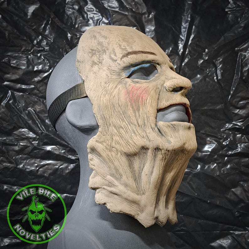 May include: A detailed, realistic, beige-colored latex mask of an elderly person with a wrinkled face. The mask has a painted-on expression of a smile with a slightly open mouth. The mask is on a mannequin head, and the mask's strap is visible.