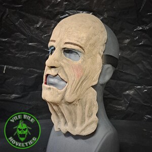 May include: A detailed, realistic, beige-colored latex mask of a man's face with a wrinkled forehead, a furrowed brow, and a wide, open mouth. The mask has a strap on the back of the head. The mask is on a gray mannequin head.