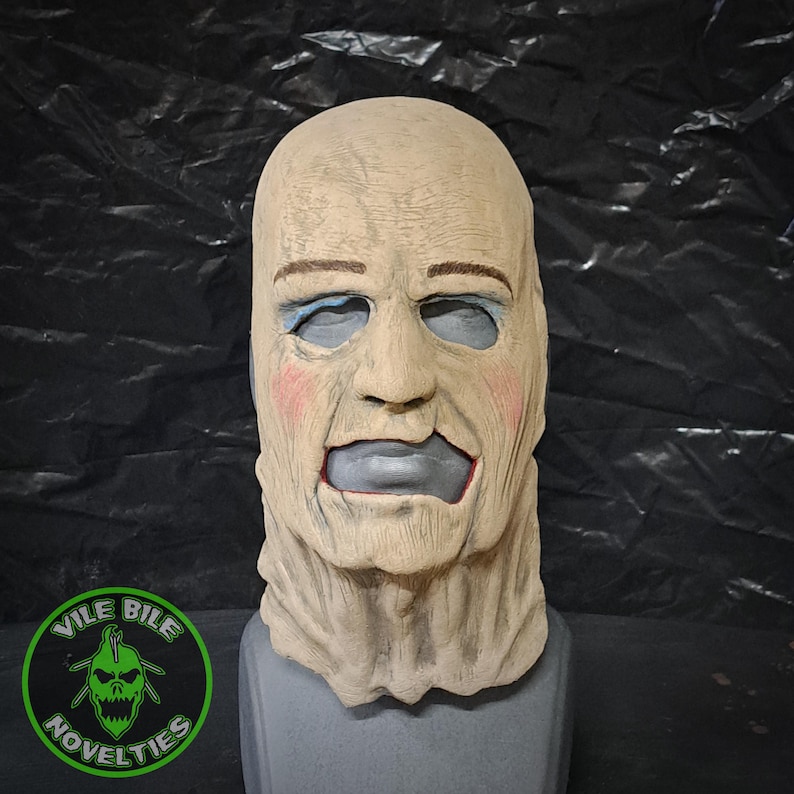May include: A beige latex mask with a bald head, painted-on eyebrows, blue eyes, and a gray mouth. The mask has a textured surface and a long neck. The mask is on a gray stand. The mask is a horror prop.