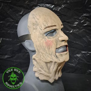 May include: A beige latex horror mask with a detailed wrinkled texture and a painted expression. The mask has a painted brown eyebrow, blue eyes, red cheeks, and a dark blue mouth. The mask is on a gray mannequin head.