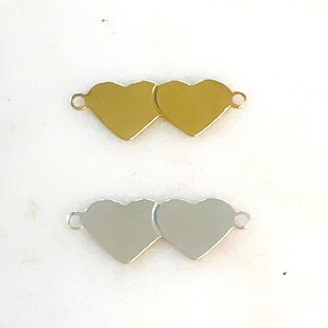 Pode incluir: Dois pingentes de metal em forma de coração, um dourado e outro prateado, com argolas de salto para prender em joias ou outras manualidades.