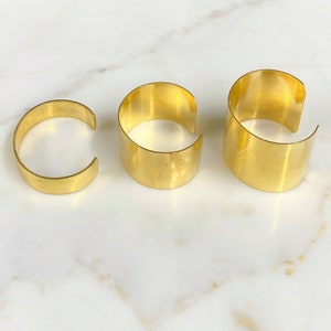 May include: Three gold-colored metal cuff bracelets of varying sizes. The bracelets have a simple, open design and a smooth, polished finish. The cuffs are arranged on a white marble surface.
