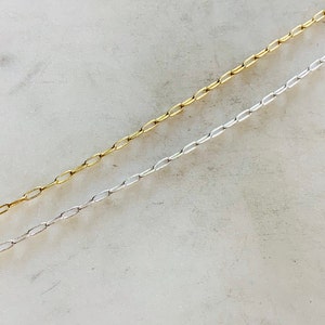K&ouml;nnte beinhalten: Zwei Ketten, eine silberne und eine goldene, liegen auf einer wei&szlig;en Oberfl&auml;che. Beide Ketten bestehen aus kleinen, ovalen Gliedern.