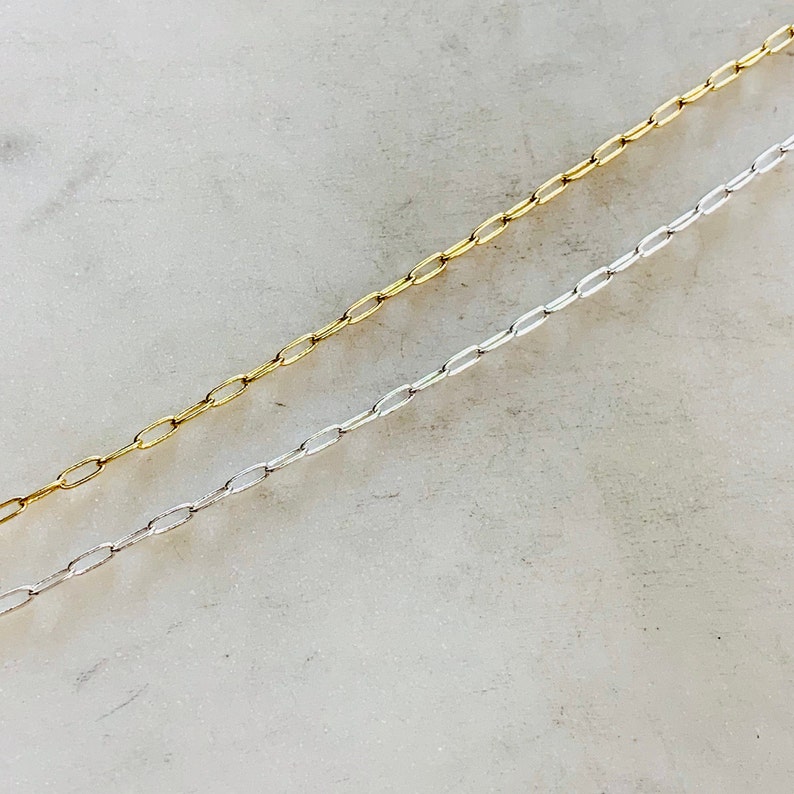 K&ouml;nnte beinhalten: Zwei Ketten, eine silberne und eine goldene, liegen auf einer wei&szlig;en Oberfl&auml;che. Beide Ketten bestehen aus kleinen, ovalen Gliedern.