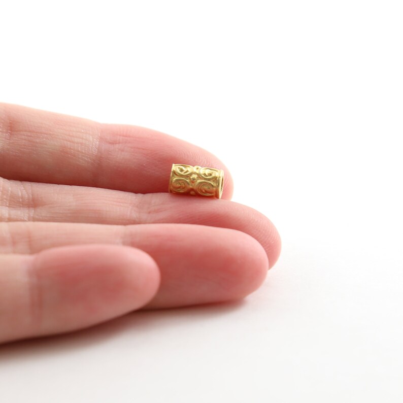 May include: A gold-colored metal bead with a decorative pattern. The bead is cylindrical in shape and has a smooth, polished finish.