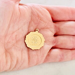 May include: Gold-toned metal charm with a detailed image of the Virgin Mary and a cross. The charm is round and has a scalloped edge.