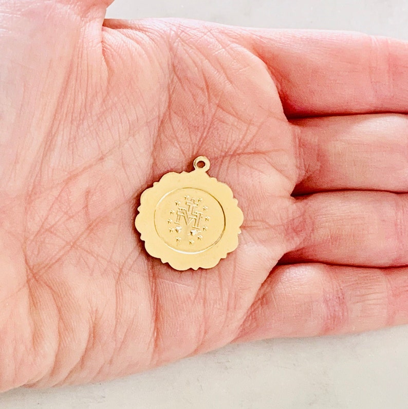 May include: Gold-toned metal charm with a detailed image of the Virgin Mary and a cross. The charm is round and has a scalloped edge.