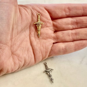 Peut inclure: Deux breloques d'&eacute;p&eacute;e orn&eacute;es, l'une dor&eacute;e et l'autre argent&eacute;e, avec des d&eacute;tails complexes. Les breloques sont pr&eacute;sent&eacute;es dans une main, ce qui permet de se faire une id&eacute;e de leur taille.