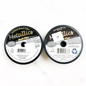 Op de afbeelding: Twee spoelen met antiek messingkleurige metalen flexdraad. De spoel links is gemarkeerd met "Fijn" en heeft een diameter van 0,014 inch en een lengte van 30 voet. De spoel rechts is gemarkeerd met "Medium" en heeft een diameter van 0,019 inch en een lengte van 30 voet.