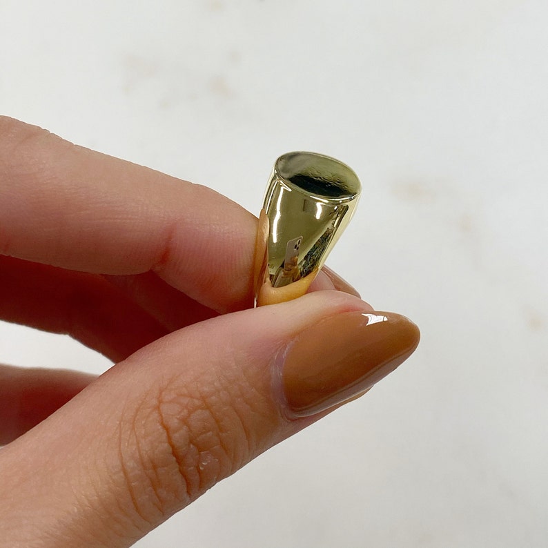 May include: A gold-colored signet ring with a smooth, oval face. The ring is held by a person with a light skin tone. The ring's surface reflects light, highlighting its polished finish. The ring is a classic jewelry piece.