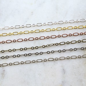 May include: Five different chain styles in silver, gold, copper, bronze, and black metal. The chains are laid out horizontally on a white surface.