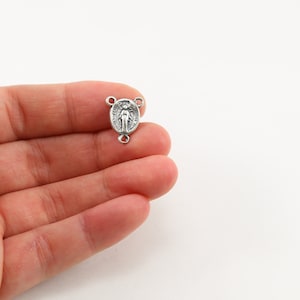 May include: Silver-toned metal rosary charm featuring a depiction of the Virgin Mary. The charm has a loop on each side for attaching to a rosary.