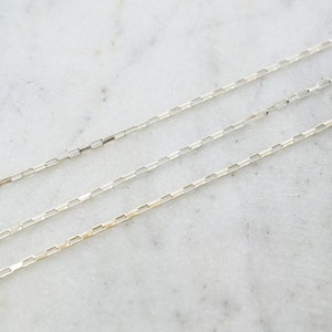 May include: Three delicate silver chain necklaces with rectangular links are displayed against a white marble surface. The chains are positioned diagonally across the frame, showcasing their fine craftsmanship and minimalist design. The chains are ideal for layering.