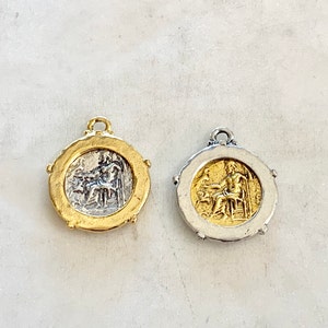 May include: Two round metal charms featuring a raised image of a person seated on a throne. One charm is gold with a silver image, and the other is silver with a gold image.
