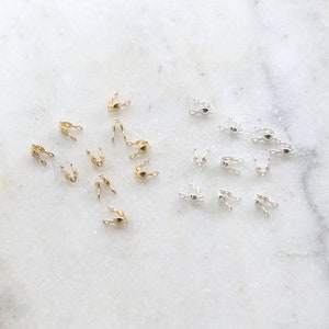 May include: A close-up of two groups of small, metal crimp beads. One group is gold and the other is silver.