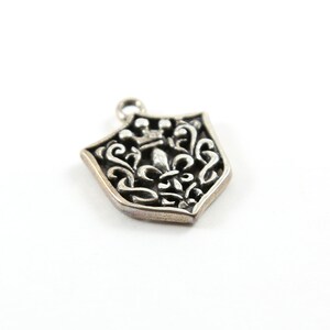 Peut inclure: Charme de pendentif en argent en forme de bouclier avec un motif de fleur de lys et une couronne au sommet.