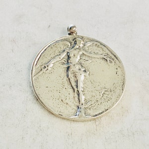 May include: A silver pendant featuring a raised relief design of a stylized human figure with outstretched arms.