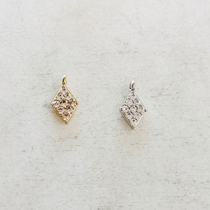 May include: Two diamond-shaped pendants, one gold-toned and one silver-toned, each encrusted with small, clear gemstones. The pendants are displayed on a light-colored, textured surface, showcasing their sparkle.