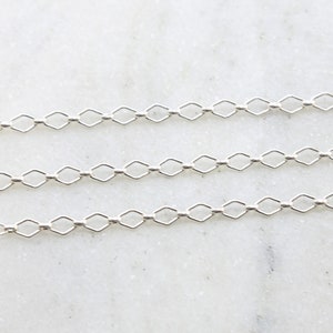 May include: Silver chain with a delicate, square-shaped link pattern. The chain is shown in three rows on a white marble surface.